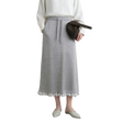 A model wearing a light gray knit midi skirt with white pom-pom trim at the hem, styled with a white blouse, a dark brown crescent bag, and off-white pointed flats. The AMII brand logo is visible in the top left corner.
