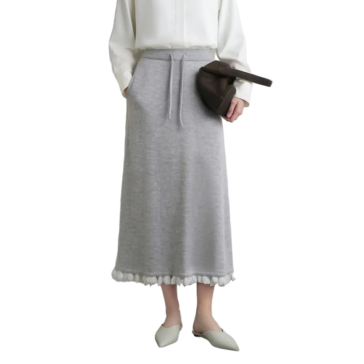 A model wearing a light gray knit midi skirt with white pom-pom trim at the hem, styled with a white blouse, a dark brown crescent bag, and off-white pointed flats. The AMII brand logo is visible in the top left corner.