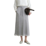 A model wearing a light gray knit midi skirt with white pom-pom trim at the hem, styled with a white blouse, a dark brown crescent bag, and off-white pointed flats. The AMII brand logo is visible in the top left corner.