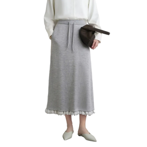 A model wearing a light gray knit midi skirt with white pom-pom trim at the hem, styled with a white blouse, a dark brown crescent bag, and off-white pointed flats. The AMII brand logo is visible in the top left corner.