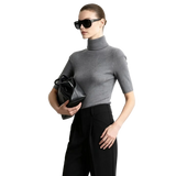 A woman modeling a fitted short-sleeve gray turtleneck sweater with black trousers and black sunglasses, holding a black handbag against a light gray studio background. The AMII brand name appears in the top left, along with a '100% sheep wool' label.