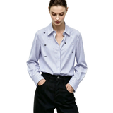A woman wearing a light blue and white vertically striped long-sleeve button-up shirt with small embroidered floral details on the chest, paired with dark trousers. The AMII brand logo is visible in the top left corner.
