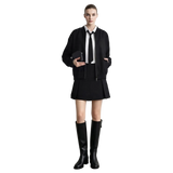 A model stands against a light gray wall wearing a black zip-front jacket over a white collared shirt with a black tie, a black pleated mini skirt, and tall black knee-high boots.