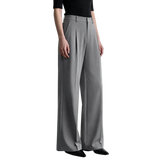 A model wearing a pair of high-waisted, wide-leg gray trousers with pleated detailing, paired with a black short-sleeve top and black pointed-toe flats. The AMII brand logo is visible in the top-left corner against a minimalist light gray studio background.