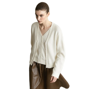 A model wearing an AMII cream-colored V-neck knit cardigan sweater and brown leather pants, standing against a minimalist light gray wall and concrete floor.