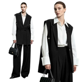 Two models wearing a black belted vest over a white long-sleeve shirt and black wide-leg trousers, standing against a light gray studio background. One model holds a black quilted handbag with a chain strap.
