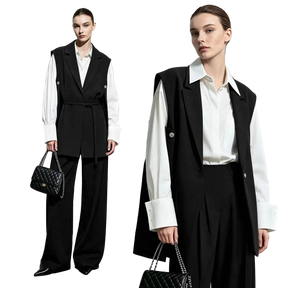 Two models wearing a black belted vest over a white long-sleeve shirt and black wide-leg trousers, standing against a light gray studio background. One model holds a black quilted handbag with a chain strap.