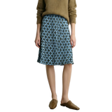 A model wearing a blue and gray geometric patterned midi skirt with a brown knit sweater and brown mule loafers, standing against a light gray wall. The brand name AMII is visible in the top left corner.