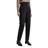 A model wearing black high-waisted, straight-leg trousers with pleats, paired with a beige sweater and black pointed-toe ankle boots. The AMII brand logo is visible in the top-left corner against a neutral light gray studio background.