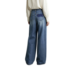 A model from behind wearing AMII wide-leg blue denim jeans with a contrasting dark blue patch pocket on the back, paired with a cream-colored blouse against a minimalist studio background.