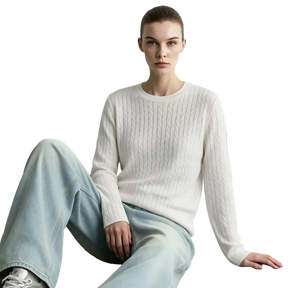A model wearing a cream cable-knit long-sleeve sweater and light-wash wide-leg jeans, sitting against a light gray wall. The brand name 'AMII' is printed in black in the top-left corner.