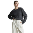 A woman wearing a dark charcoal gray long-sleeve knit sweater and cream-colored high-waisted trousers, standing against a light gray studio background with the AMII logo in the top left corner.