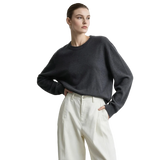 A woman wearing a dark charcoal gray long-sleeve knit sweater and cream-colored high-waisted trousers, standing against a light gray studio background with the AMII logo in the top left corner.