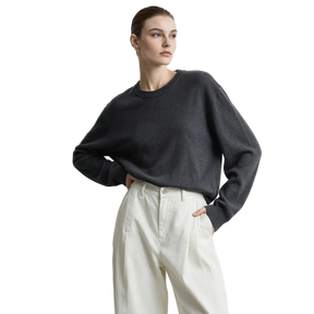 A woman wearing a dark charcoal gray long-sleeve knit sweater and cream-colored high-waisted trousers, standing against a light gray studio background with the AMII logo in the top left corner.