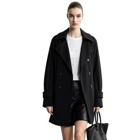 A model wearing a black double-breasted AMII coat over a white top and black shorts, holding a black textured handbag against a light gray studio background. An informational graphic on the left highlights the wrinkle-resistant, no-iron fabric.
