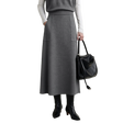 A model wearing a gray A-line midi skirt with two front patch pockets, styled with a gray top, white long-sleeve shirt, black ankle boots, and holding a black leather shoulder bag. The AMII brand logo is visible in the top left corner.
