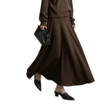 A model wearing a brown knit long-sleeve top and matching high-waisted A-line midi skirt, styled with black pointed-toe mule heels and a black leather handbag. The AMII brand logo is visible in the top left corner against a neutral gray studio background.