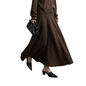 A model wearing a brown knit long-sleeve top and matching high-waisted A-line midi skirt, styled with black pointed-toe mule heels and a black leather handbag. The AMII brand logo is visible in the top left corner against a neutral gray studio background.