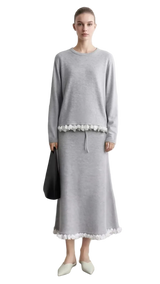 A model wears a light gray long-sleeve midi dress with white floral embroidery at the waist and hem, standing against a light gray wall. The left side of the image features a text overlay panel with the brand name 'AMII' and a close-up of the white lace embroidery.