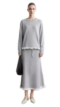 A model wears a light gray long-sleeve midi dress with white floral embroidery at the waist and hem, standing against a light gray wall. The left side of the image features a text overlay panel with the brand name 'AMII' and a close-up of the white lace embroidery.